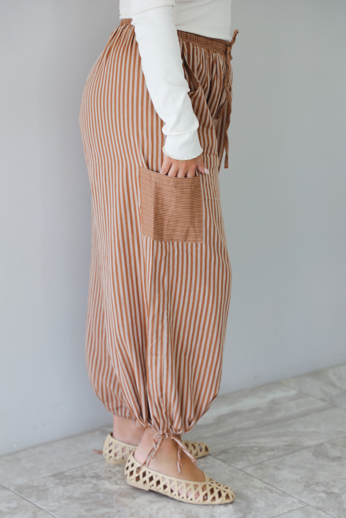 A sideview and closeup of a woman wearing a pair of camel and cream striped balloon pants that have an elastic and drawstring waistline with drawstring details at the bottom hem at the ankles, standing indoors against a white wall with white marble flooring. 