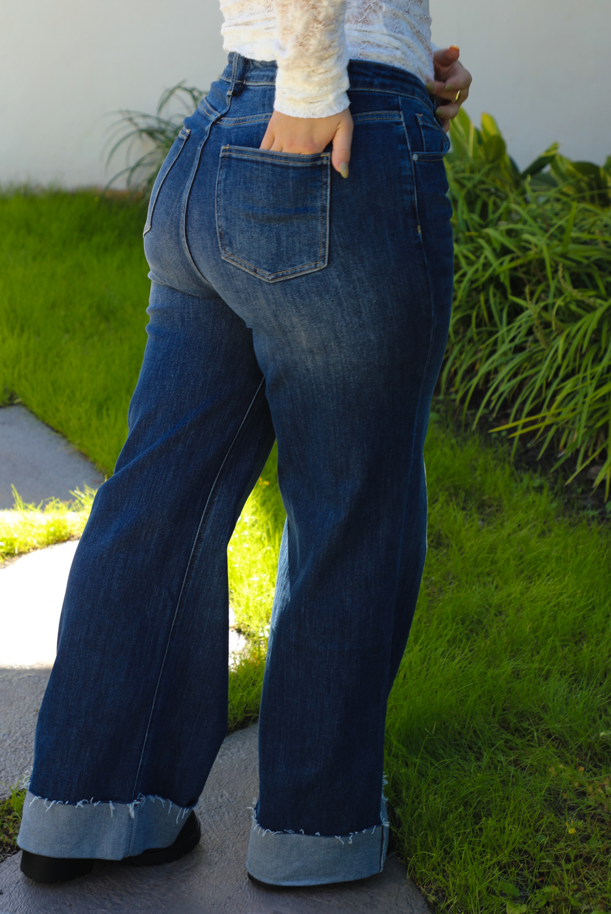 Back of a woman model wearing dark denim blue jeans outdoors. They have a high waistline, functional button and zipper closures in the front, functional front and back pockets, and a slightly flared cut with a cuffed hem. 