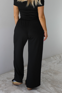 Woman wearing a two piece set featuring an off the shoulder top and high waisted pants. The photo shows a closeup of the back of the pants.  The photo is taken indoors against a white wall with white marble flooring.
