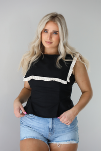 Woman wearing a black sleeveless top with a square-cut bib that has a cream colored pleated detail around the bib. The top is paired with light-wash denim shorts. The photo is taken indoors against a white wall.