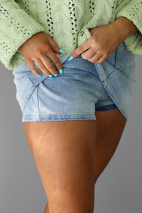 Woman wearing a light wash denim skort that has a high waisted, a button closure that's off center, a zipper closure, with shorts underneath. The photo is taken indoors against a white wall and shows a closeup of the shorts under the skirt. 