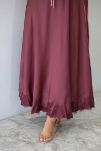 A woman modeling a long, burgundy maxi dress with short puff sleeves, a lace-up sweetheart neckline, and a flowing satin-like skirt, focusing in on the lace hem of the dress, standing indoors against a white wall with white marble flooring. 