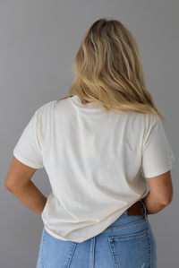 Woman wearing a white t-shirt with text and graphics showing two kitties with the writing 'show me your kitties', untucked to show the full length of the back of the top. The photo is taken indoors against a white wall.
