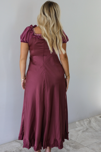 A woman shown from behind modeling a long, burgundy maxi dress with short puff sleeves, zipper closure in the back, shown in front of a white wall with white marble flooring. 