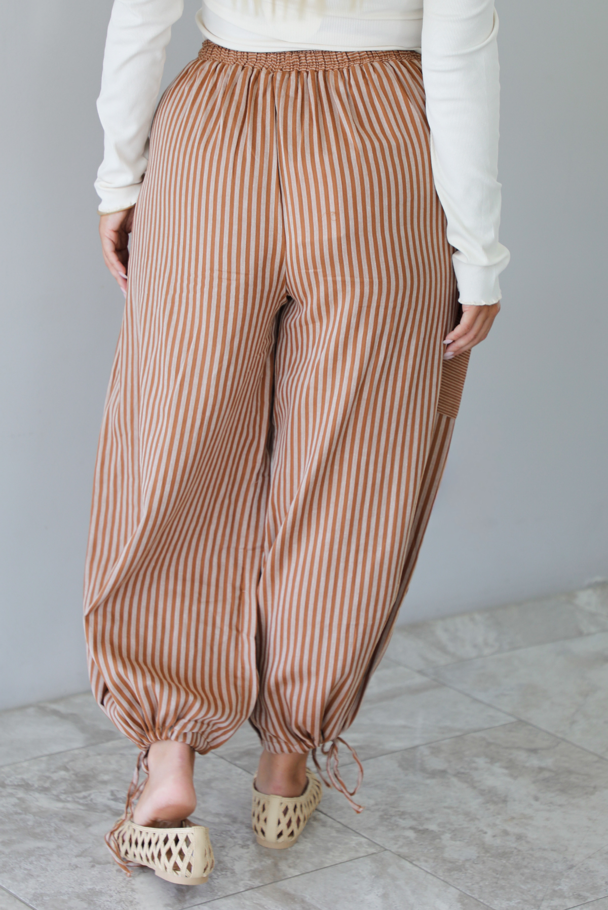 The back and a closeup of a woman wearing a pair of camel and cream striped balloon pants that have an elastic and drawstring waistline with drawstring details at the bottom hem at the ankles, standing indoors against a white wall with white marble flooring. 