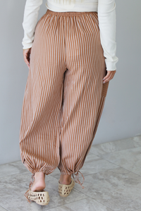 The back and a closeup of a woman wearing a pair of camel and cream striped balloon pants that have an elastic and drawstring waistline with drawstring details at the bottom hem at the ankles, standing indoors against a white wall with white marble flooring. 