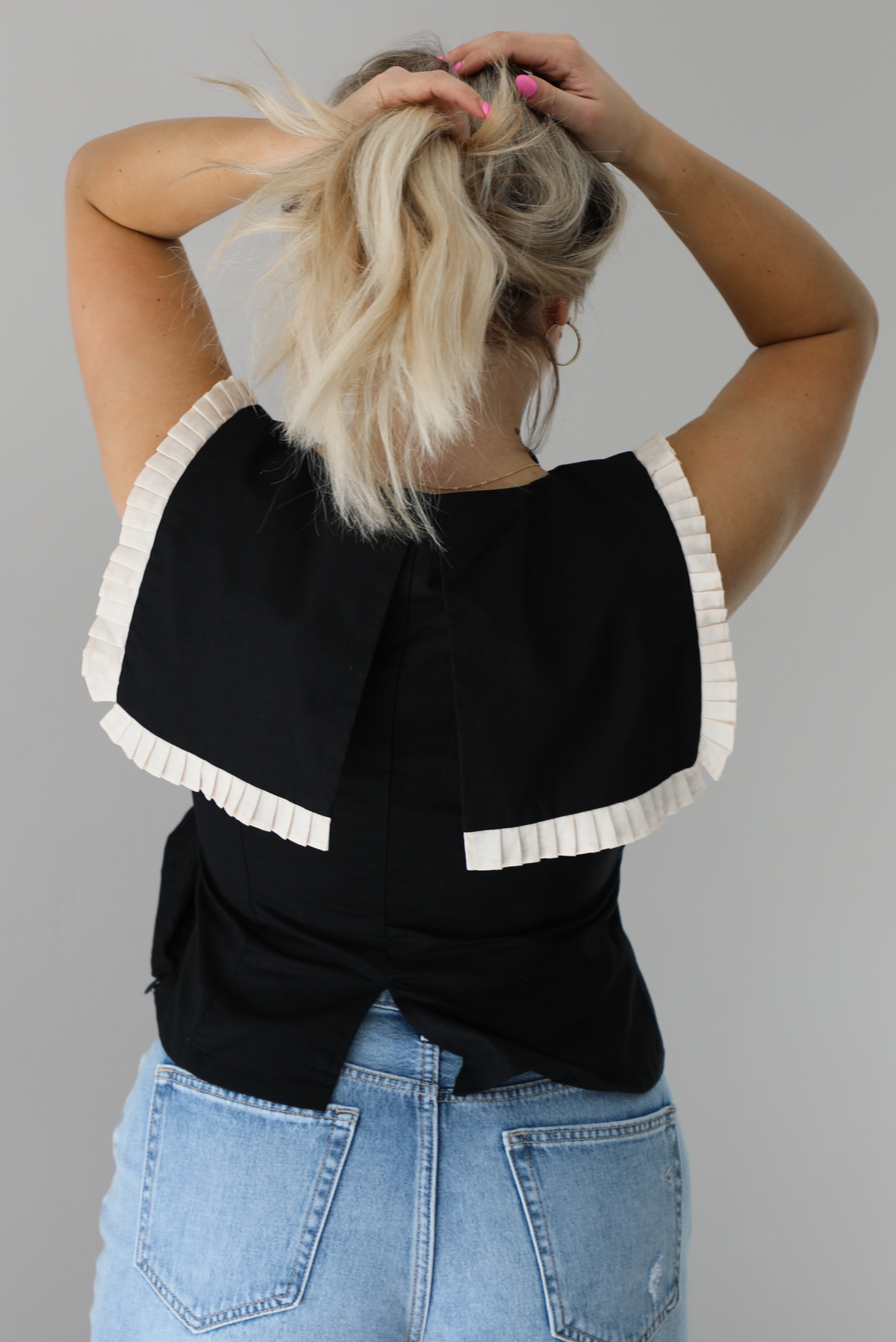 Woman wearing a black sleeveless top with a square-cut bib that has a cream colored pleated detail around the bib. The top is paired with light-wash denim shorts. The photo shows the back of the top. The photo is taken indoors against a white wall.