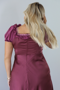 A woman shown from behind modeling a long, burgundy maxi dress with short puff sleeves, zipper closure in the back, shown in front of a white wall.
