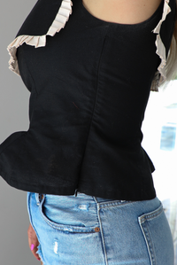 Woman wearing a black sleeveless top with a square-cut bib that has a cream colored pleated detail around the bib. The top is paired with light-wash denim shorts. The photo shows the side of the top, which has a zipper closure. The photo is taken indoors against a white wall.