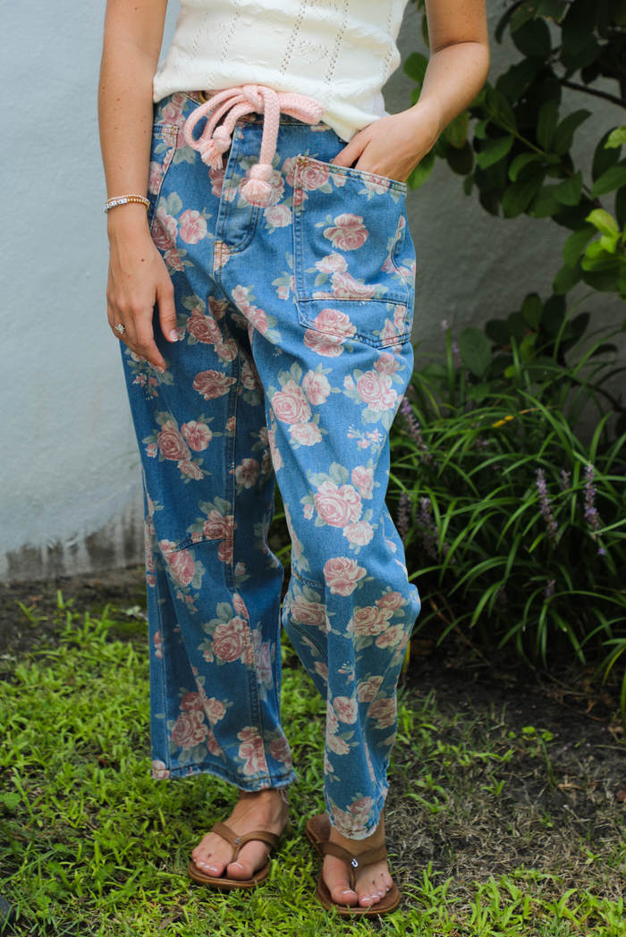 Woman wearing blue denim jeans that have a pink, white, and green rose print pattern throughout The jeans have a high waistline, functional front and back pockets, a tie closure at the waist, and a barrel cut. The photo is taken outdoors with greenery in the back.