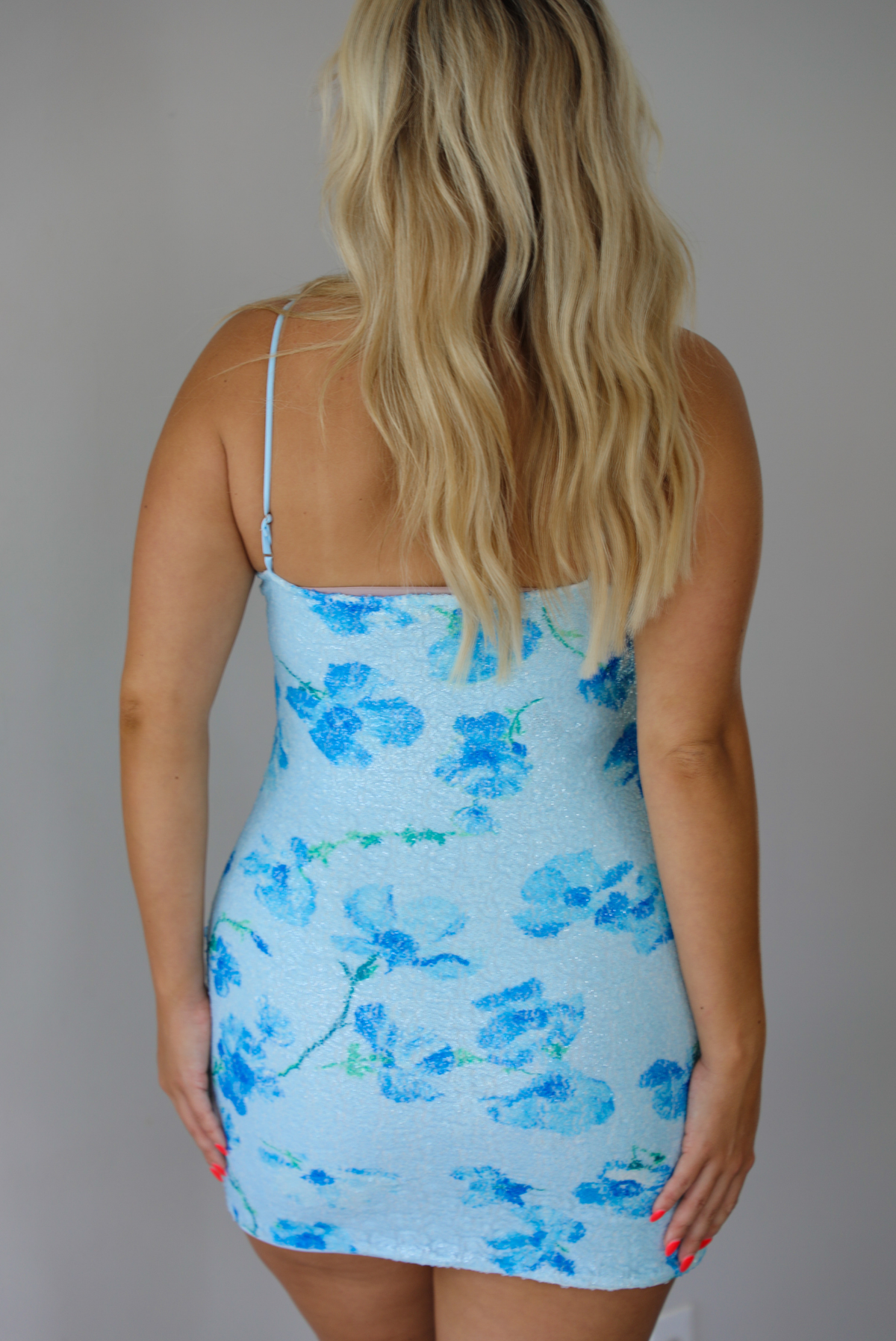 A blonde woman modeling a strapless light blue bodycon mini dress with a darker blue floral pattern, paired with brown flat sandals, standing indoors against a white wall. The photo shows the back of the dress with its adjustable straps.