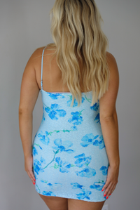 A blonde woman modeling a strapless light blue bodycon mini dress with a darker blue floral pattern, paired with brown flat sandals, standing indoors against a white wall. The photo shows the back of the dress with its adjustable straps.