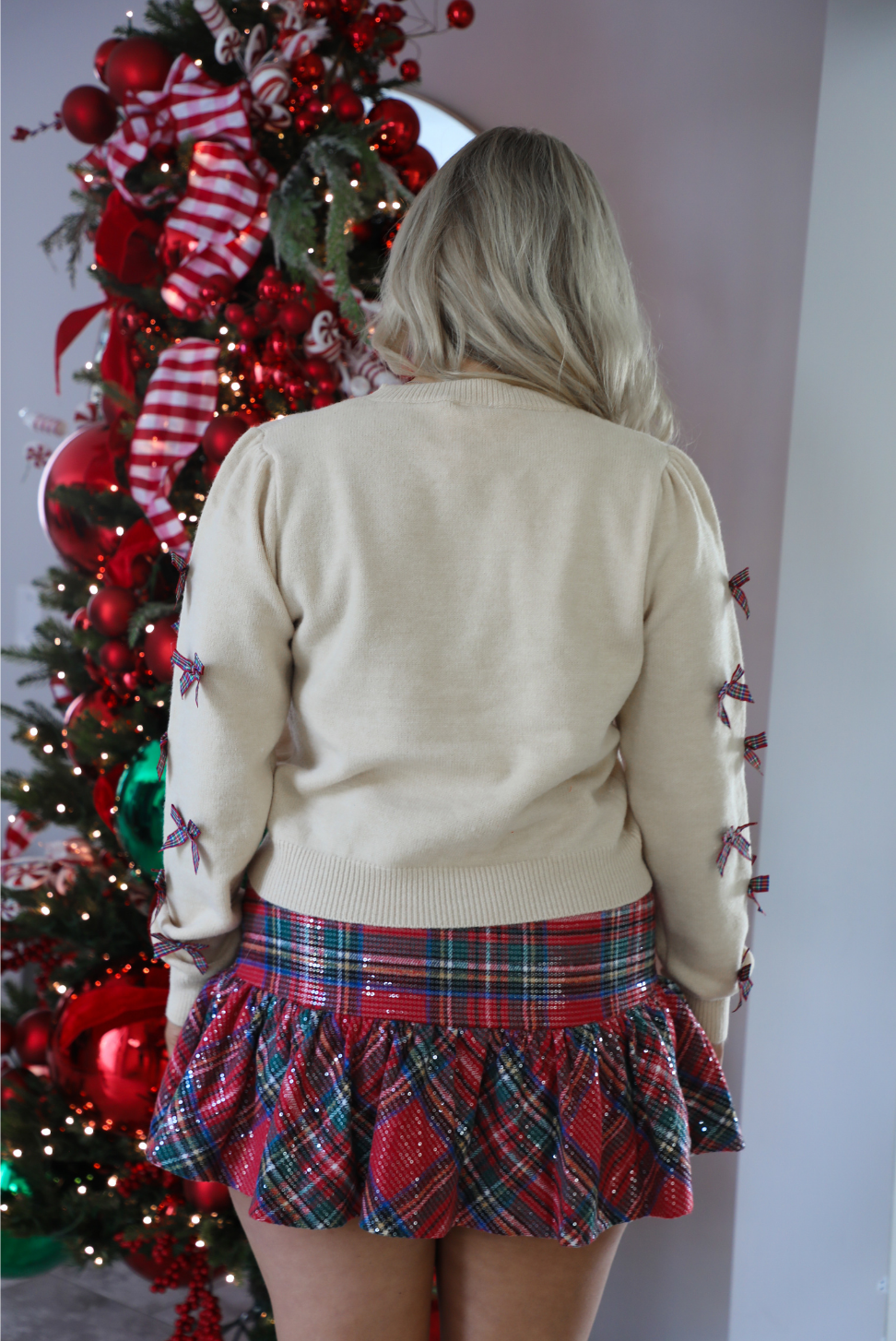 Back of a woman model wearing a sand colored sweater with multi-colored plaid bow details on the sleeves with a multicolored plaid print skirt. The photo showcases the full length of the sweater, which stops at the upper hip on the 5'7" model. The photo is taken inside a modern home with a decorated Christmas tree in the back.