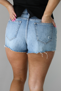 Woman wearing a light-wash pair of denim shorts that have distressed detailing. The photo shows a closeup of the shorts with the models right hand in the back pocket. The photo is taken indoors against a white wall. 