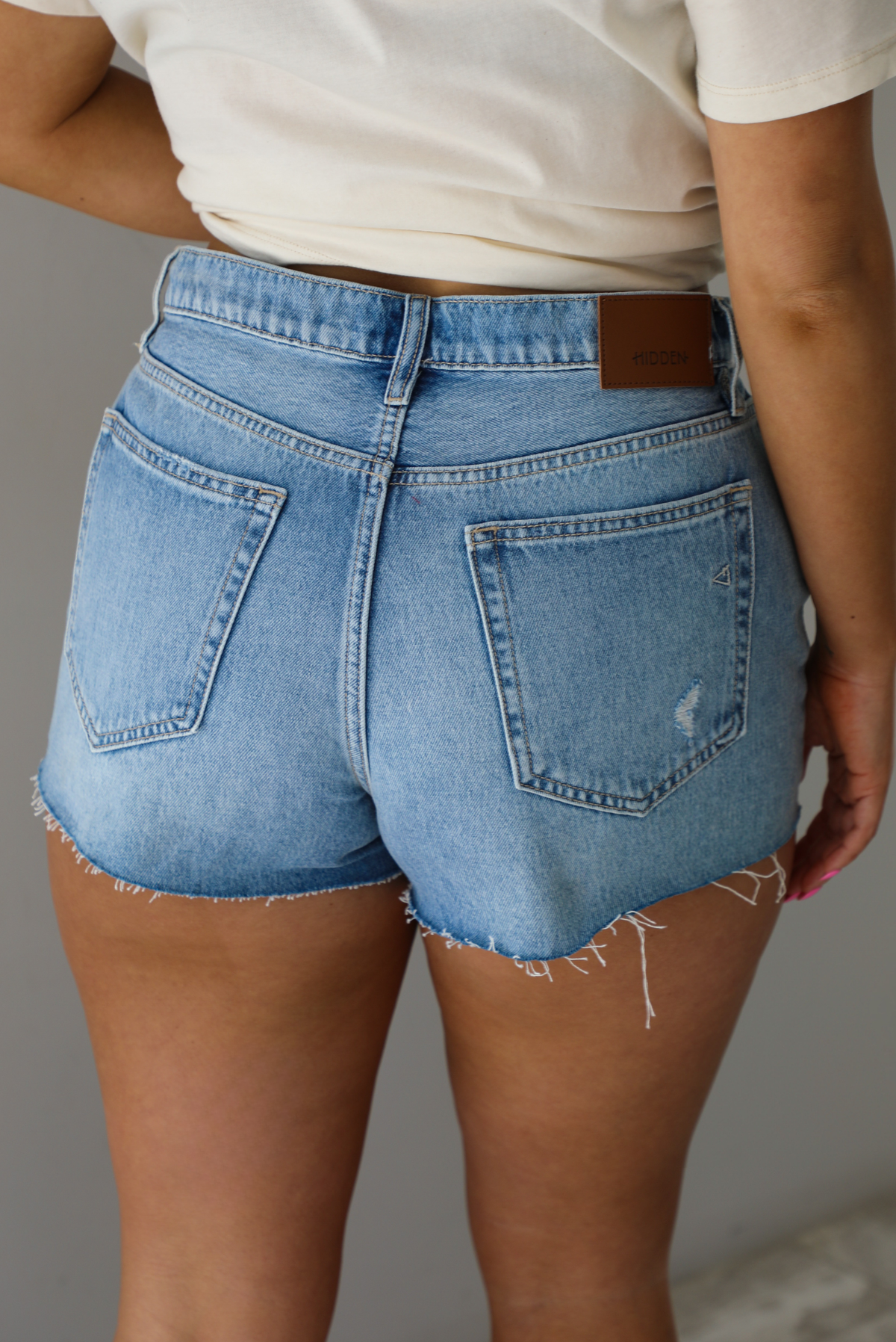 Woman wearing a light-wash pair of denim shorts that have distressed detailing. The photo shows the back of the shorts. The photo is taken indoors against a white wall.