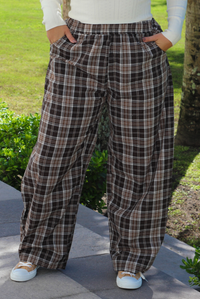 A woman shown from the waist down wearing a cream colored long sleeved top paired with brown plaid pants, standing outdoors on a stone path with trees and greenery in the background. 