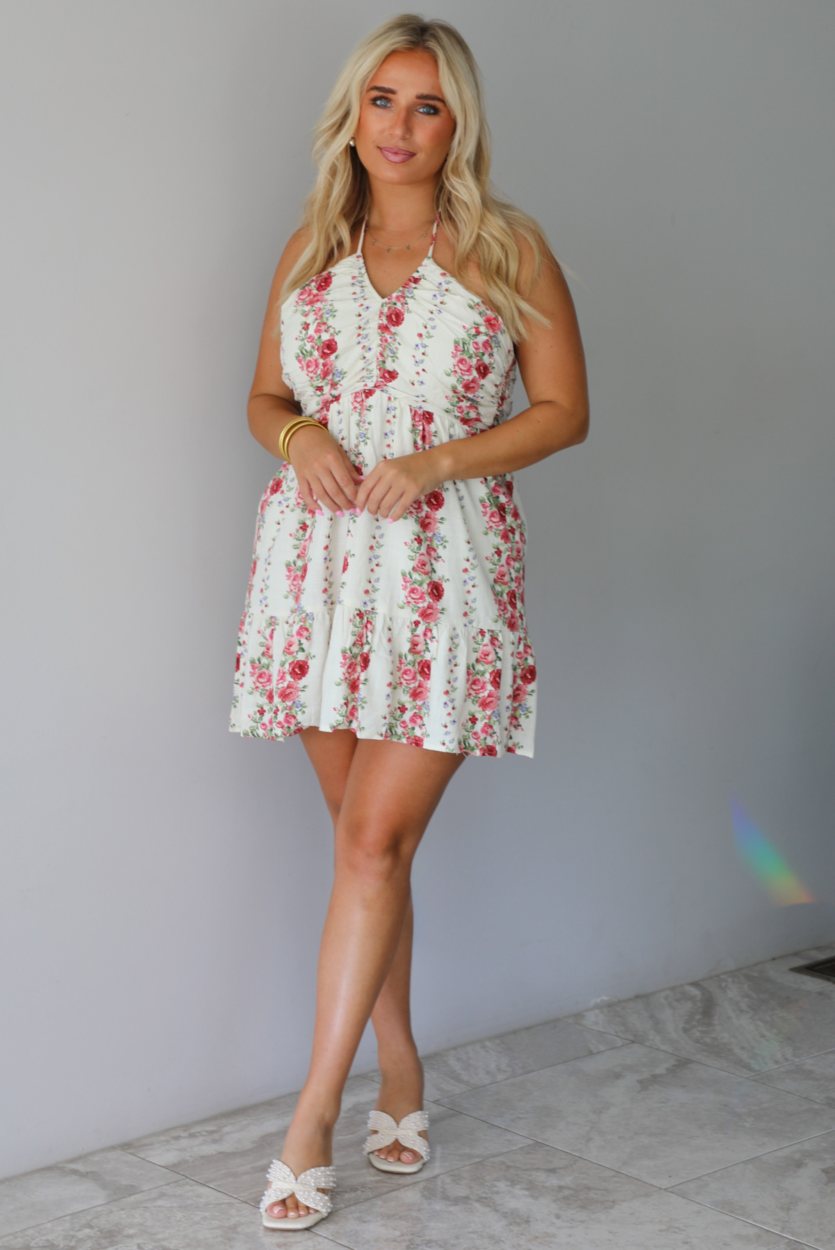 A woman wearing a white halter mini dress with a red and pink floral print, standing indoors against a white wall with white marble flooring. 