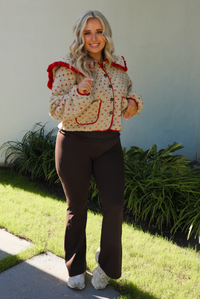 Woman model wearing a tan and red sherpa cardigan with a collared neckline and red trim with dark brown ahtleisure pants. The photo is taken outdoors with a white wall and greenery in the back. 