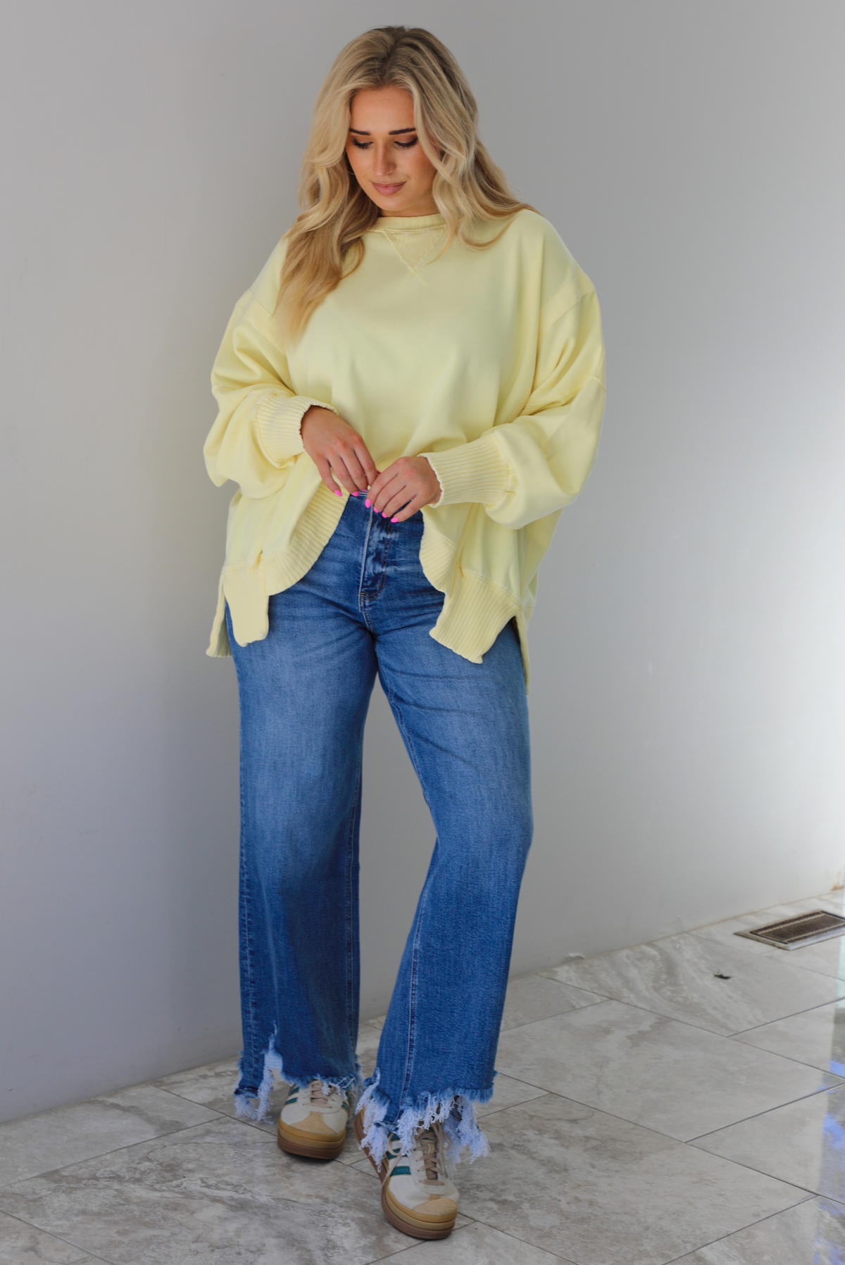 Woman wearing a pale yellow oversized sweater with dark wash denim and sneakers. The photo is taken indoors against a white wall with white marble flooring. 