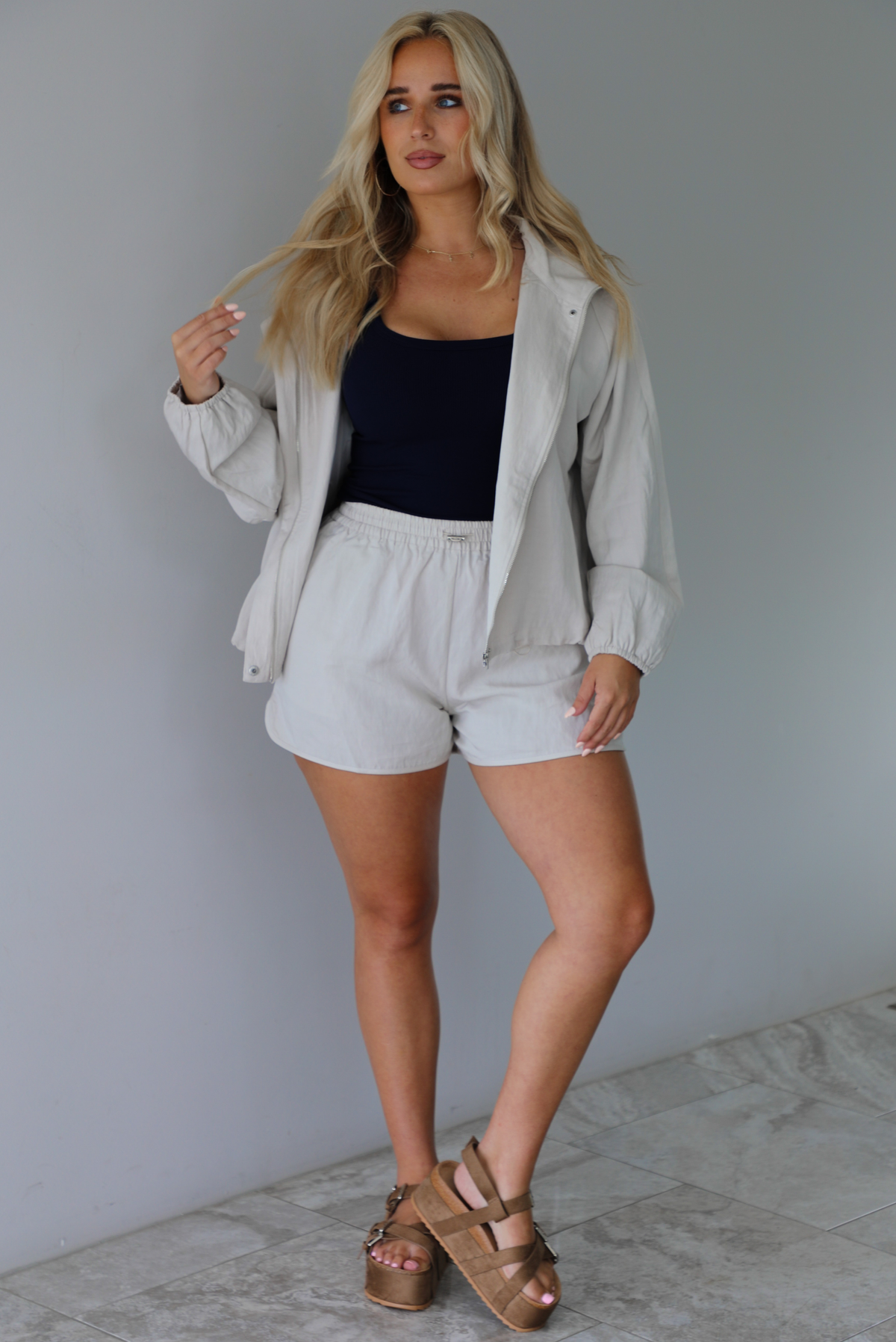 Woman wearing a stone colored two piece set featuring a zip up jacket and shorts with a navy tank tucked into the shorts indoors against a white wall with white marble flooring. 