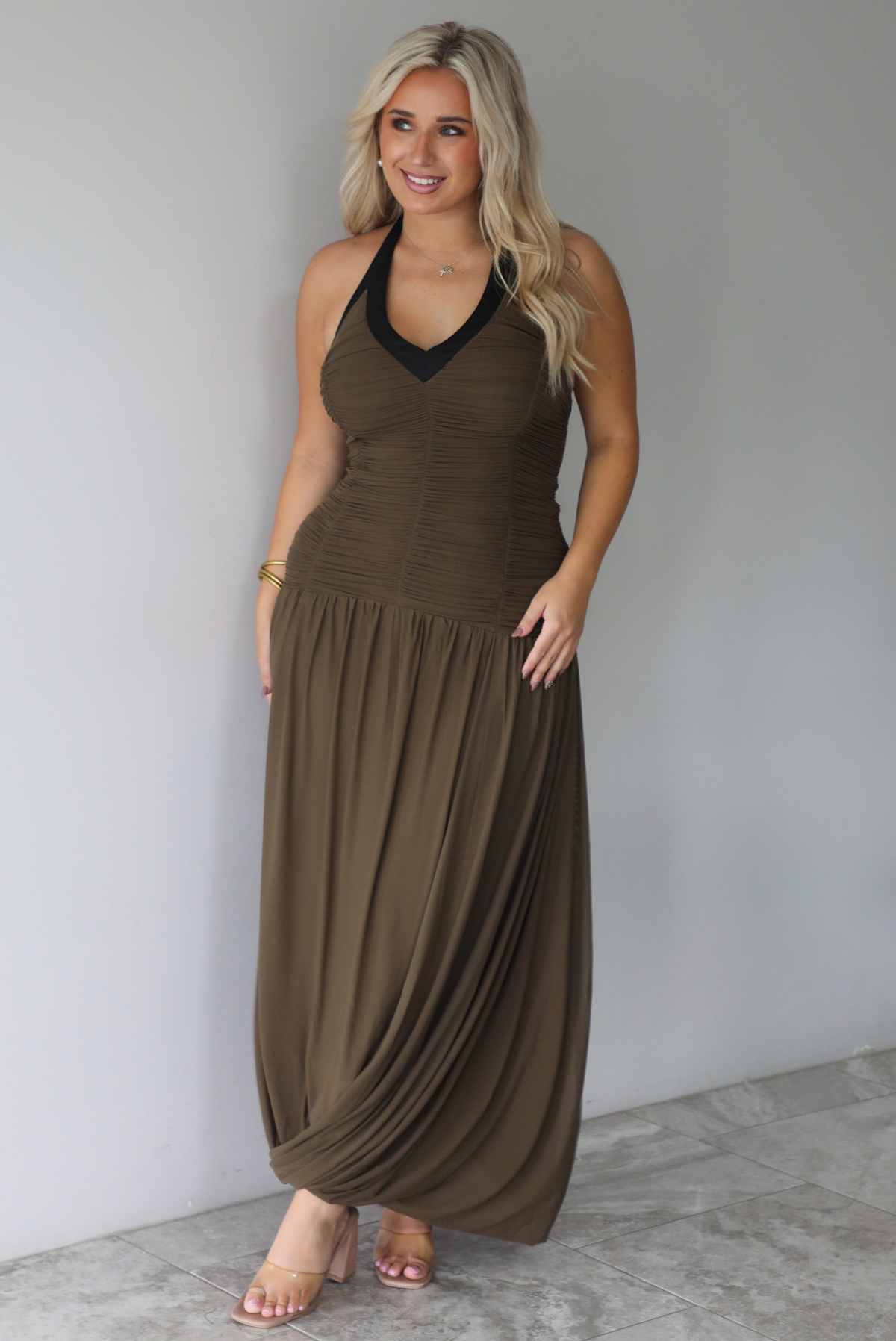 A woman wearing an olive maxi dress with black trim detailing at the v-halter neckline, has a fitted bodice and drop waist design, with a maxi-length flowy skirt, standing indoors against a white wall with white marble flooring. 