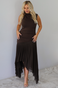 A woman wearing a brown midi dress that has a high neckline, is fitted until the upper thigh, then leads to sheer mesh that's flowy ands an asymmetrical hem, standing indoors against a white wall with white marble flooring. 