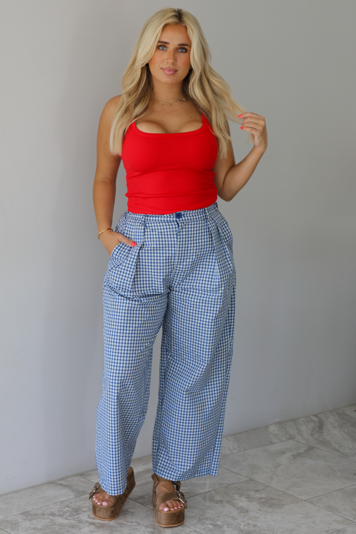 Woman wearing a red tank top high waisted navy and white plaid pants indoors against a white wall with white marble flooring. 