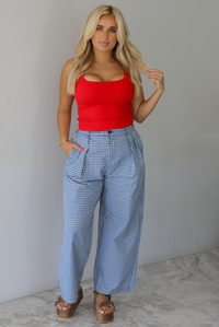 Woman wearing a red tank top high waisted navy and white plaid pants indoors against a white wall with white marble flooring. 