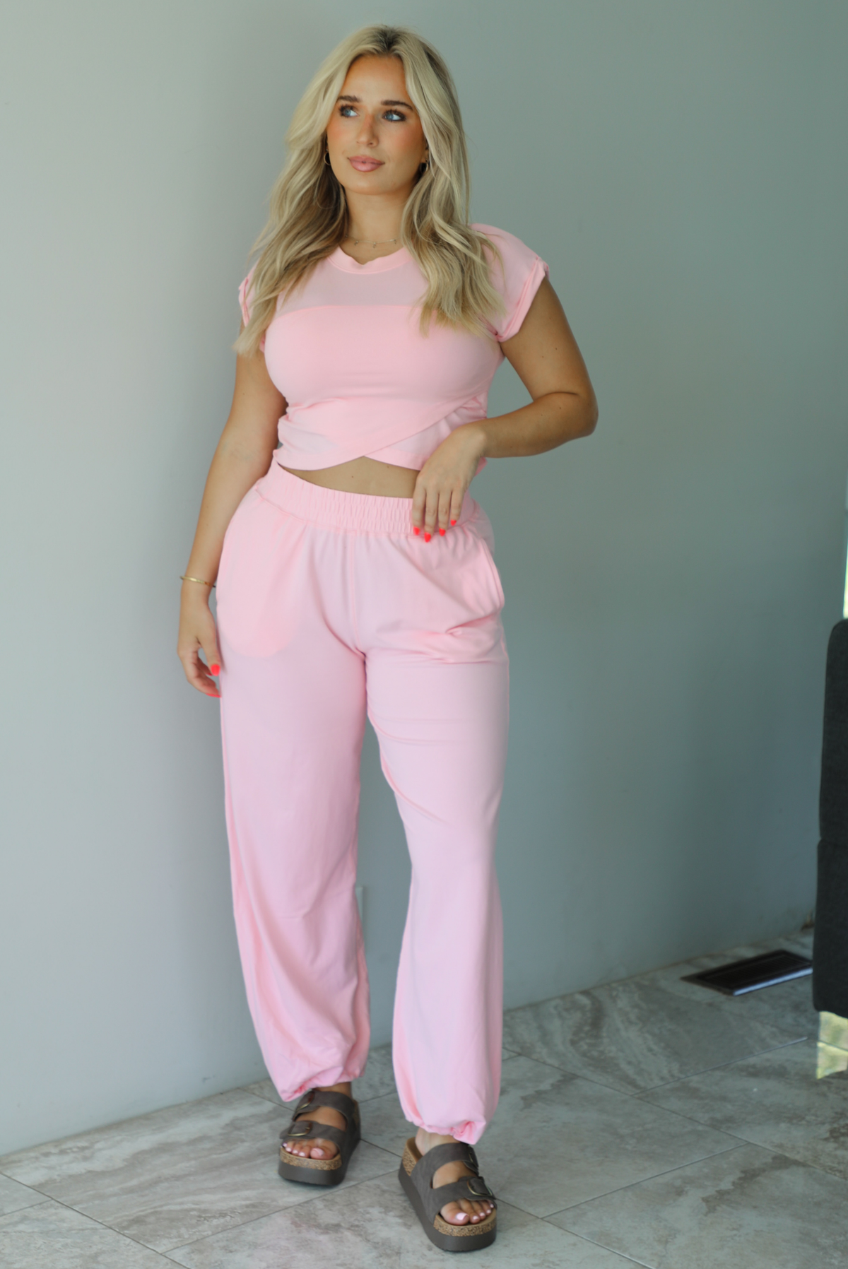 Woman wearing a pink two piece set featuring a cropped short sleeved top and high waisted jogger-style pants indoors against a white wall with white marble flooring. 