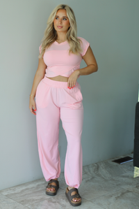 Woman wearing a pink two piece set featuring a cropped short sleeved top and high waisted jogger-style pants indoors against a white wall with white marble flooring. 