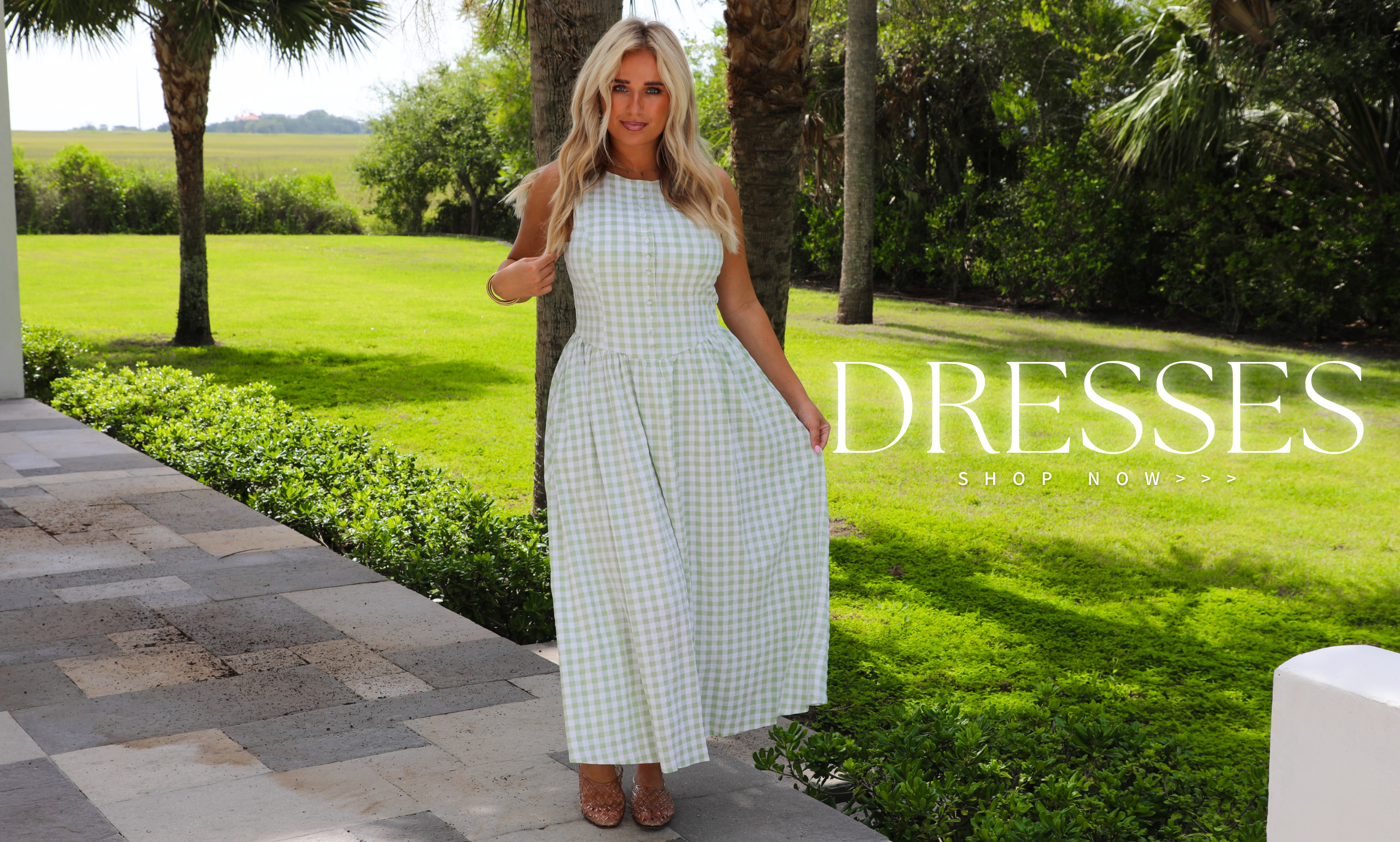 A woman modeling a sleeveless white and light green gingham maxi dress in an outdoor garden setting with palm trees and a stone walkway. A text overlay on the right reads 'DRESSES SHOP NOW'.