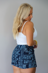 A woman shown from behind wearing a white tank tucked into a denim dark navy mini skirt with a white bandana paisley print pattern throughout, standing indoors against a white wall.
