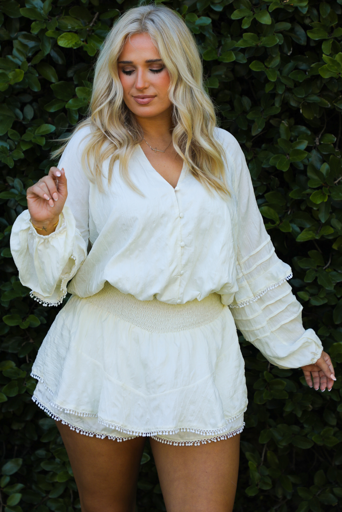 Woman model wearing an off-white romper outdoors in front of greenery. 