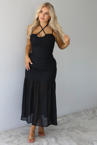 Woman wearing a black two piece set featuring a tank and high waisted, maxi-length skirt that has a sheer mesh hem. The photo is taken indoors against a white wall with white marble flooring. 