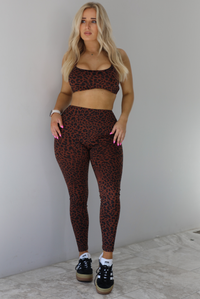 Woman wearing a brown and white leopard print two piece athleisure set that consists of a sports bra and high waisted leggings. The photo is taken indoors against a white wall with white marble flooring. 