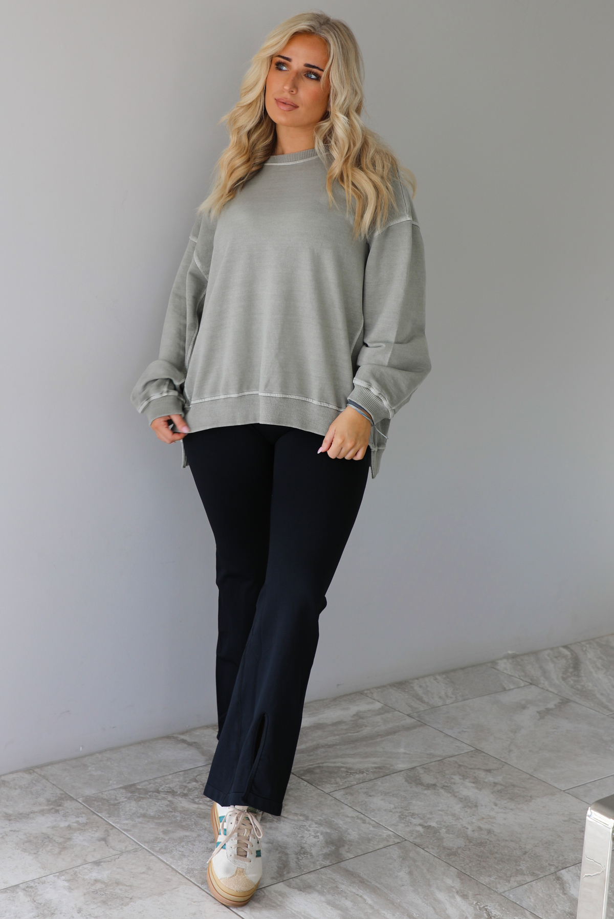 Woman wearing a tan oversized sweater with black leggings that have a slightly flared hem with split hem details in the front. The photo is taken indoors against a white wall with white marble flooring. 