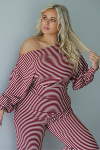 A woman wearing a maroon and white striped two piece matching set featuring a slouchy off the shoulder long sleeved top and mid-rise, folded over waistline wide leg pants, standing indoors against a white wall.