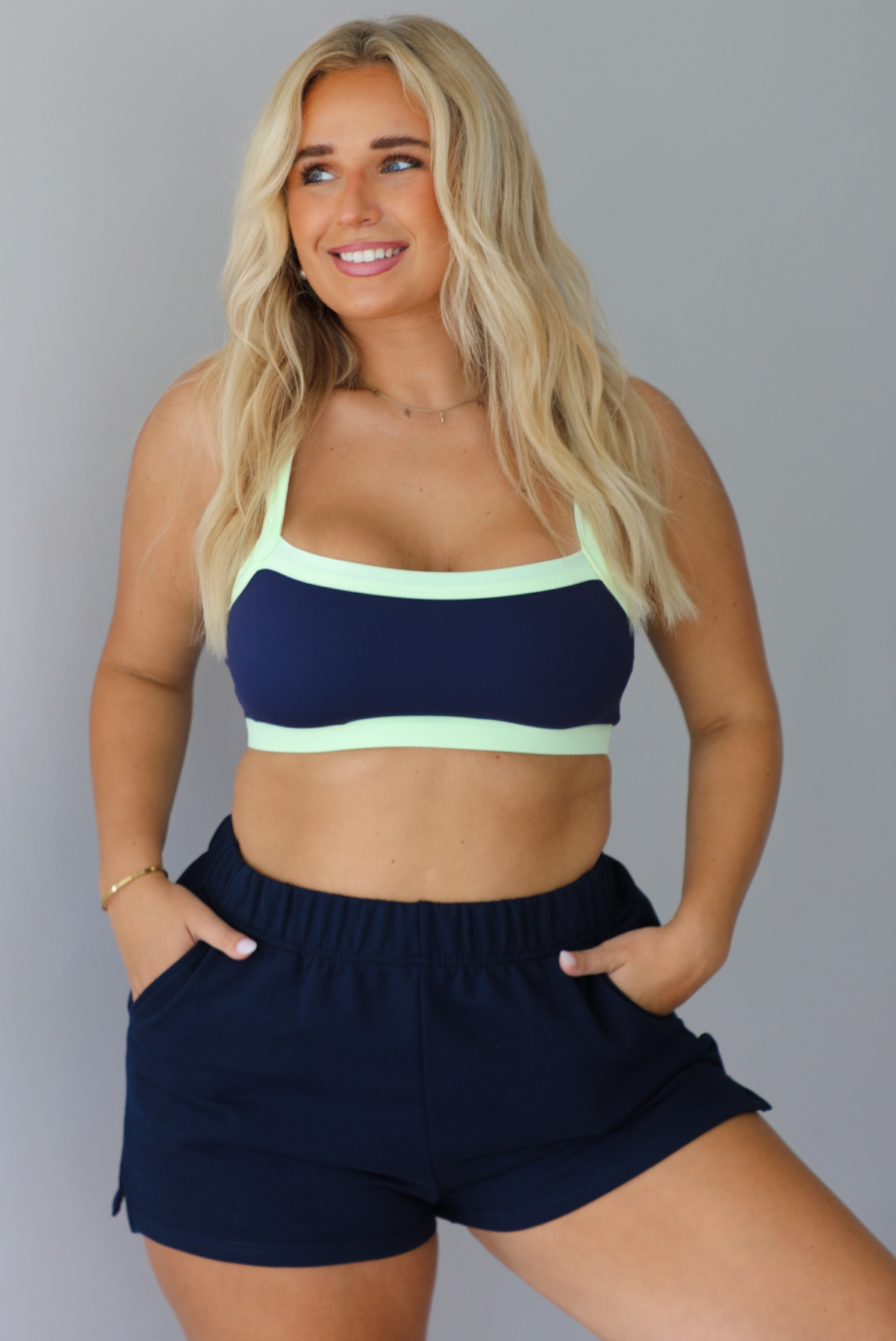 A blonde woman wearing a navy blue two-piece lounge set with light green trim, consisting of a sports bra and sweat shorts, standing indoors against a white wall.