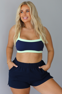 A blonde woman wearing a navy blue two-piece lounge set with light green trim, consisting of a sports bra and sweat shorts, standing indoors against a white wall.
