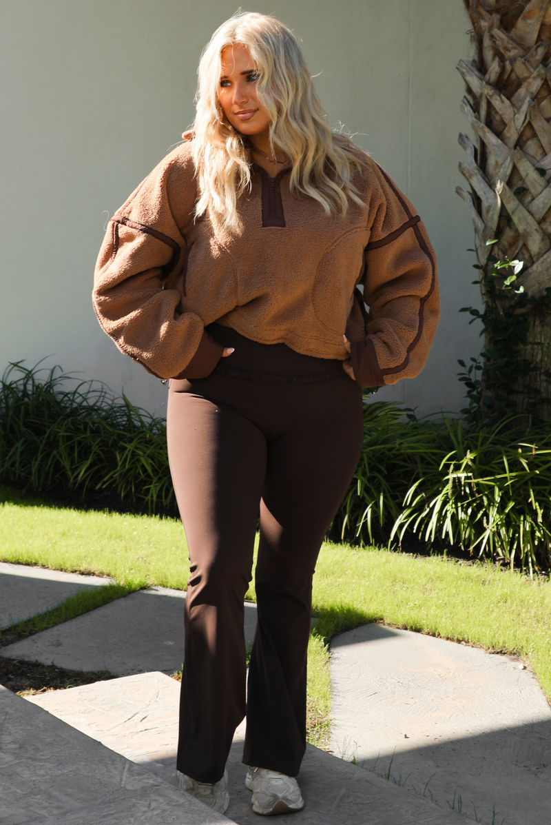 Woman model wearing a tan and brown quarter zip sweater with dark brown athleisure pants. The photo is taken outdoors against some greenery and a white wall. 