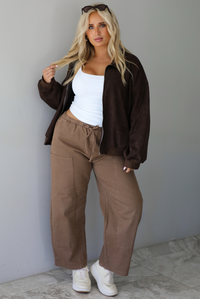 A woman wearing a dark brown suede jacket with a white tank and mocha colored mid-to-low rise pants, standing indoors against a white wall with white marble flooring.