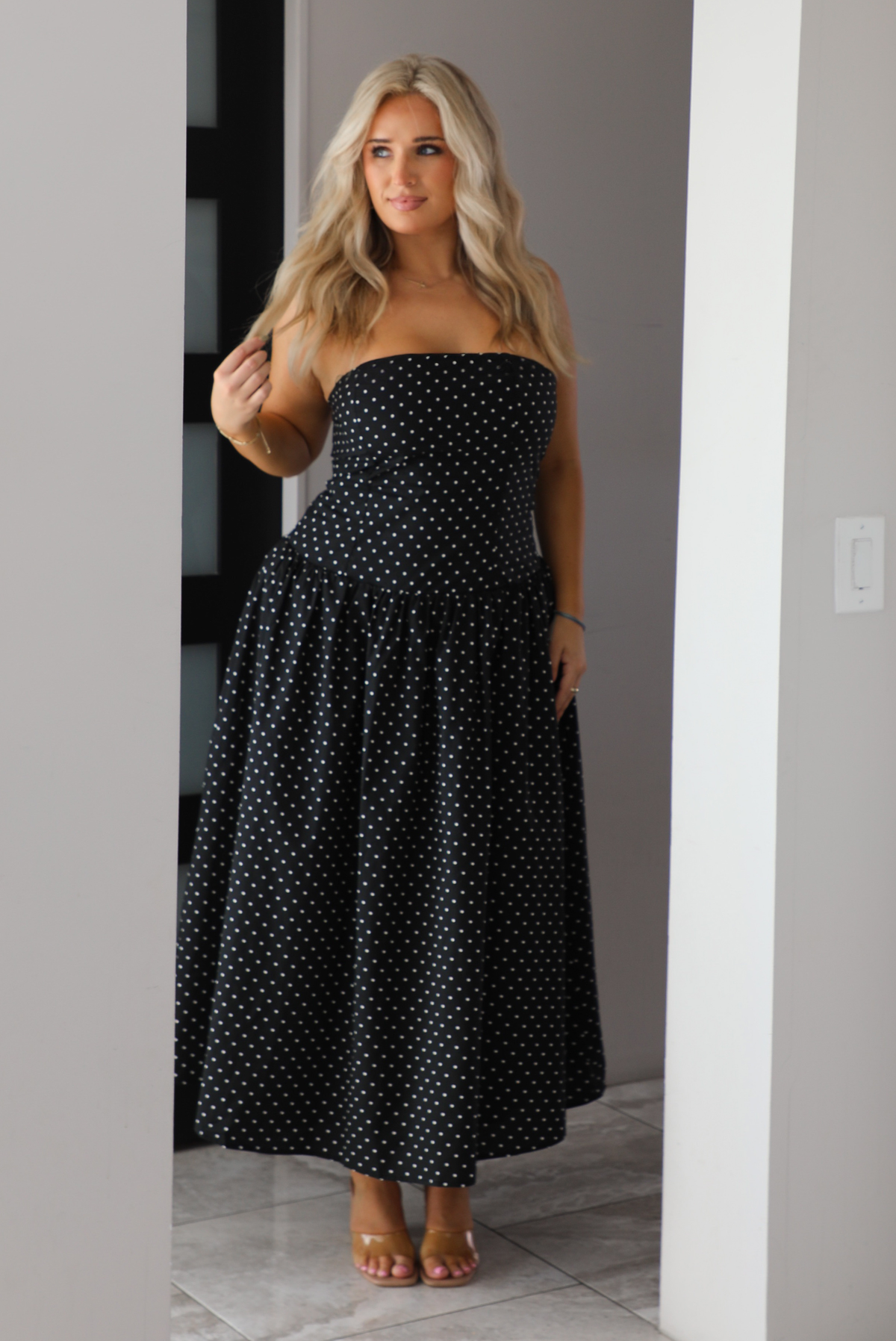 Woman model wearing a strapless/sleeveless, black with white polka dots, maxi length dress indoors. It has a fitted bodice with a flowy skirt. 