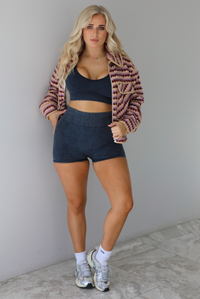 Woman wearing a pink, burgundy, lilac, and sand colored jacket with a charcoal colored two-piece set featuring a cropped sports bra and high waisted short biker shorts. The photo is taken indoors against a white wall with white marble flooring. 
