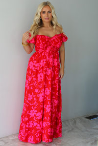 A woman modeling a bright red off-the-shoulder maxi dress with a vibrant pink floral pattern, standing indoors agains a white wall with white marble flooring. 