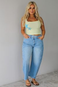 Woman wearing a yellow cropped tank with turquoise starfish studded details on the side with light wash barrel denim jeans, while standing indoors against a white wall with white marble flooring. 