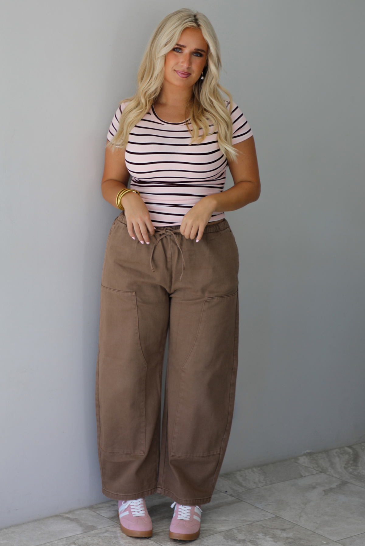 A woman wearing a pink baby tee that has a burgundy and white striped design throughout, paired with mocha barrel denim, standing indoors against a white wall with white marble flooring. 