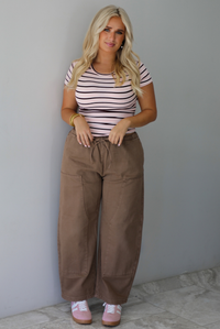 A woman wearing a pink baby tee that has a burgundy and white striped design throughout, paired with mocha barrel denim, standing indoors against a white wall with white marble flooring. 