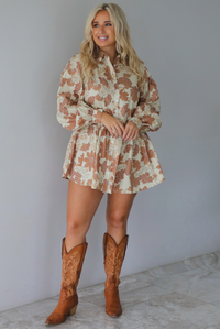 A woman wearing a cream dress with a tan fall floral print pattern throughout, collared neckline, functional button closures along the front, long sleeves that cinch tighter at the wrist, and a slightly flowy skirt, standing indoors against a white wall with white marble flooring. 