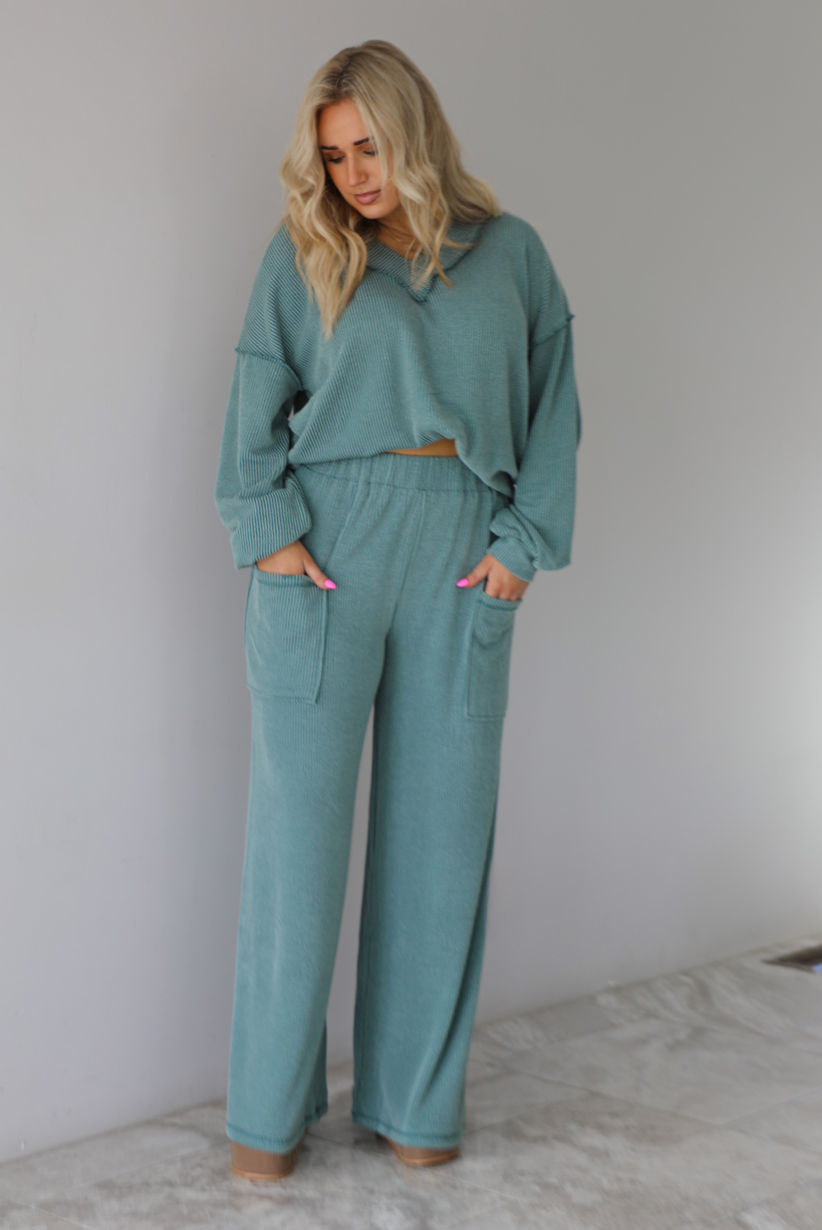 Woman wearing a teal two piece set that has a v-neck long sleeved oversized top and high waisted pants. The top is tucked into the woman's bra to give it a cropped look. The photo is taken indoors against a white wall with white marble flooring. 