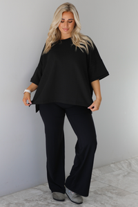 Woman wearing a black oversized box tee with longer short sleeves, a high-low design, and split hem details on either side. The tee is paired with black flared leggings. The photo is taken indoors against a white wall with white marble flooring. 